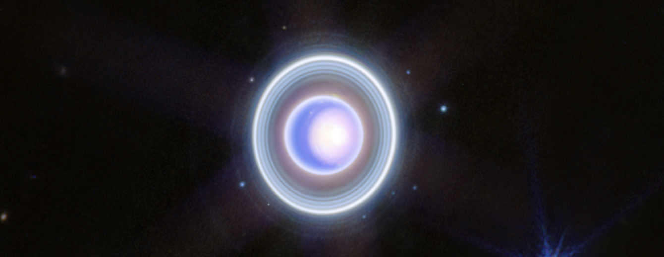 The planet Uranus on a black background. The planet appears blue with a large, white patch taking up the right half. The patch is whitest at the center, then fades into blue at it expands from right to left. A thin outline of Uranus is also white. Around the planet is a system of nested rings. The outermost ring is the brightest while the innermost ring is the faintest. Unlike Saturn’s horizontal rings, the rings of Uranus are vertical and so they appear to surround the planet in an oval shape. There are 9 blueish white dots scattered around the rings.