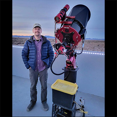 Photograph of a person standing on a concrete patio wearing a hat and a blue coat next to a large black-and-red telescope. In the background is a desert landscape, with mountains in the distance.