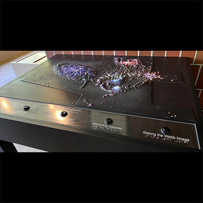 A table with a tactile display of Stephan’s Quintet on top takes up most of the frame. It appears to be sitting in an open room, against a red brick wall. The galaxies vary in terms of texture, elevation of certain characteristics, and colors. The galaxy on the left is mostly dark purple, and the texture resembles puffy, clump-like material. Toward the back of the table, there is splotchy red, vein-like material. Toward the front, there are two smooth areas of light blue and purple material surrounded by a half-circle ridge of bumpy, gray material. There are labeled buttons across the front of the table, but only two can be read. The second from right, “Interacting Galaxies,” and far right, “Making the Webb Image.” Braille runs below each button’s label.