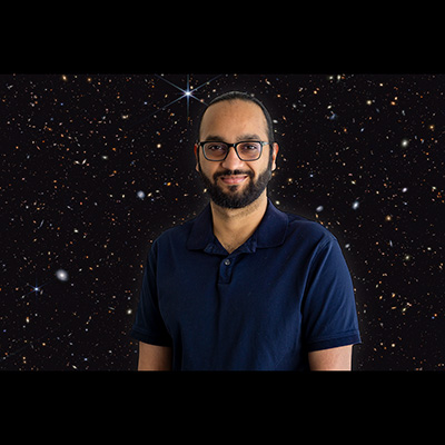 Gentleman with black hair, mustache, beard, and glasses, and wearing a dark blue, short-sleeved shirt, against a dark sky field featuring a multicolored sea of galaxies.