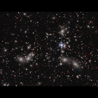 A crowded galaxy field on a black background, with one large star dominating the image just right of center. Three areas are concentrated with larger white hazy blobs on the left, lower right, and upper right above the single star. Scattered between these areas are many smaller sources of light; some also have a hazy white glow, while many other are red or orange. Even without zooming in, different galaxy shapes are detectable, like spirals, ovals, and arcs.