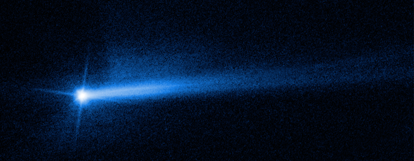 A bright blue spot is at the left-center of the image, which has a black background. The spot is the Didymos-Dimorphos system after impact from the DART spacecraft. The center bright spot has 3 diffraction spikes extending from its core at the 1 o’clock, 7 o’clock, and 10 o’clock positions. There is a small amount of dusty haze just below the southern pole of the center dot. Two tails of ejecta that appear as white streams of material extend out from the center at the 2 o’clock and 3 o’clock positions.