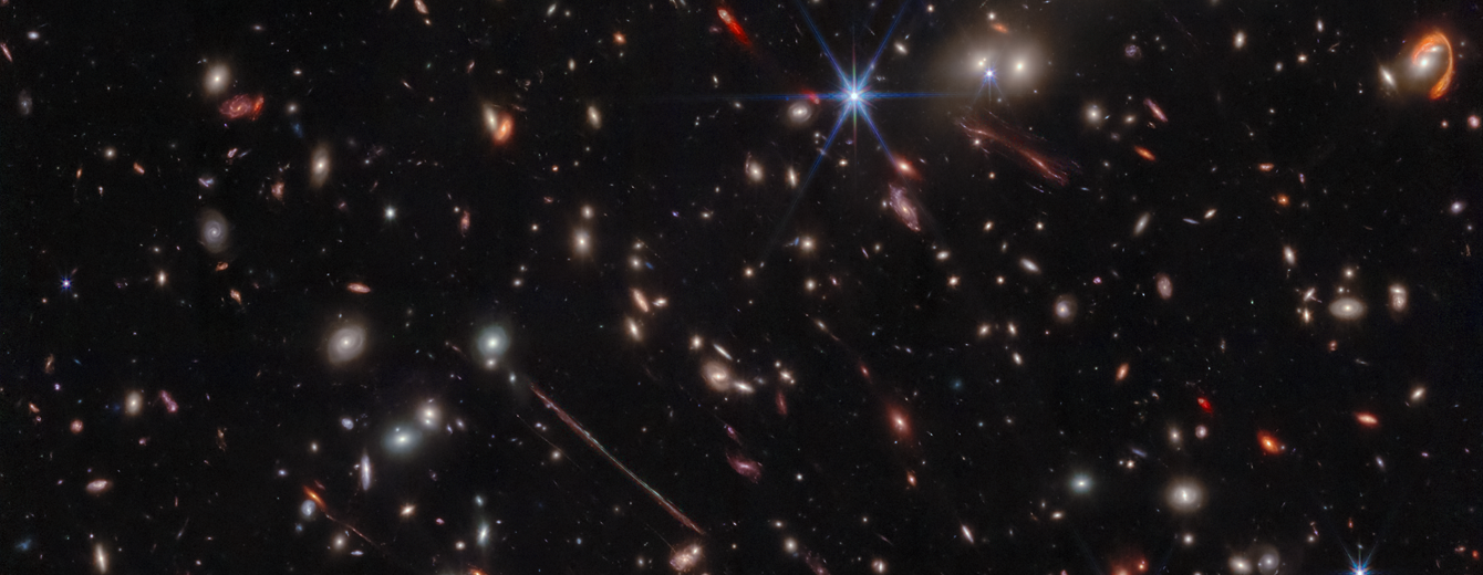 A black background is scattered with hundreds of small galaxies of different shapes, ranging in color from white to yellow to red. Some galaxies are distorted, appearing to be stretched out or mirror imaged. Near the center, a particularly long and thin line stretches from ten o’clock to four o’clock. At upper right, a red swoosh extends about three-quarters of the way around a pair of galaxies. A handful of foreground stars display eight diffraction spikes.