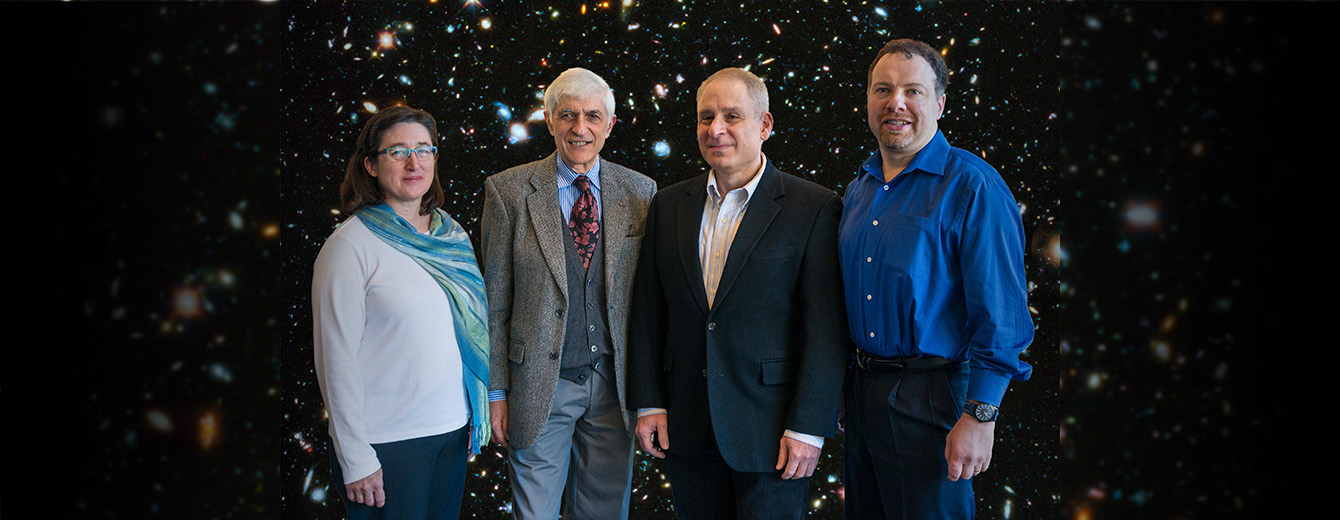 Photograph of 4 prize-winners from left to right, Susana Deustua, Nino Panagia, Andy Fruchter, Adam Riess. In the background is an image of the Hubble Deep Field, a field of colorful galaxies in the darkness of the cosmos.
