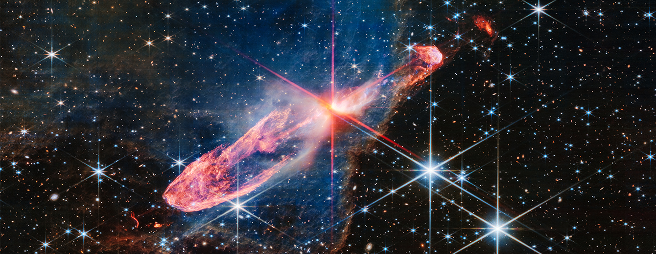At the center is a thin horizontal orange cloud known as Herbig-Haro 46/47 that is uneven with rounded ends, and tilted from bottom left to top right. It takes up about two-thirds of the length of this angle, but is thin at the opposite angle. At its center is a red-and-pink star with prominent, eight-pointed diffraction spikes. It has a central yellow-white blob. The orange lobe to the left is fatter. Just off the edge is a tiny red arc that curves in the opposite direction. The right lobe is thinner, and ends in a smaller orange semi-circle that has a faint purple outline. Just off the edge of this lobe is a slightly smaller orange sponge-like blob. A thin, undulating blue line runs from the central stars through the right lobe. A delicate, semi-transparent blue cloud known as a nebula drifts toward the top of the image and peters out toward the left of the frame, but toward the right and bottom, it ends in a soft ridge set off in a translucent orange. The background is filled with stars and galaxies.