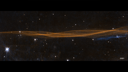 A horizontal, undulating linear structure resembling overlapping strings of yarn extends edge-to-edge across the frame, but takes up only a quarter of the width at its widest point. Mostly orange with some blue, these are filaments of glowing gasses, which resemble lines in a wrinkled bedsheet. The WFPC2 image observation date "2001" is in the bottom right corner.