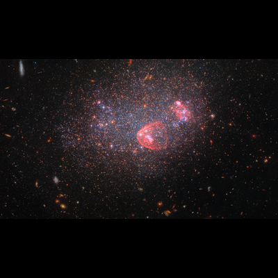 A collection of stars and galaxies fill the scene against a dark background. The image is dominated by a dense collection of stars which make up the irregular galaxy UGC 8091. The stars span a variety of colors, including blue and orange, with patches of blue occupying the central part of the galaxy. There are also visible circular regions of red/pink gas within the galaxy.