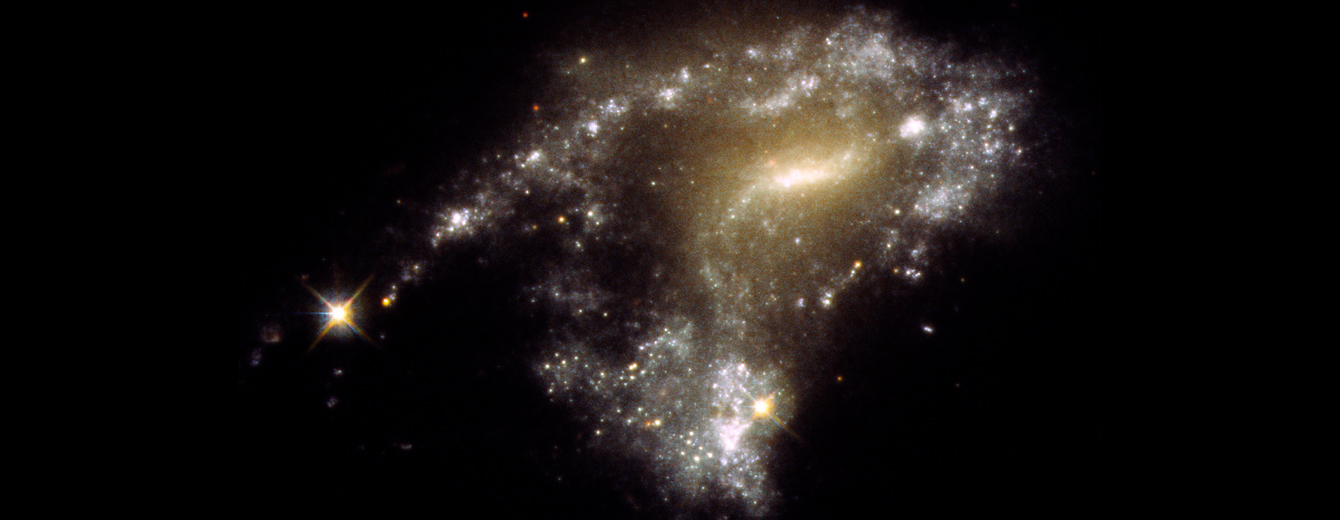 This is a picture of a galaxy with a peculiar S-shape. It has a bright milky-white core at the center. Twin arms of blue stars wrap around the core. One arm looks particularly stretched out due to the gravitational tidal pull of a neighboring galaxy. Bright, young, whitish star clusters are strung along the arm like a string of pearls. They formed as a result of the collision process.