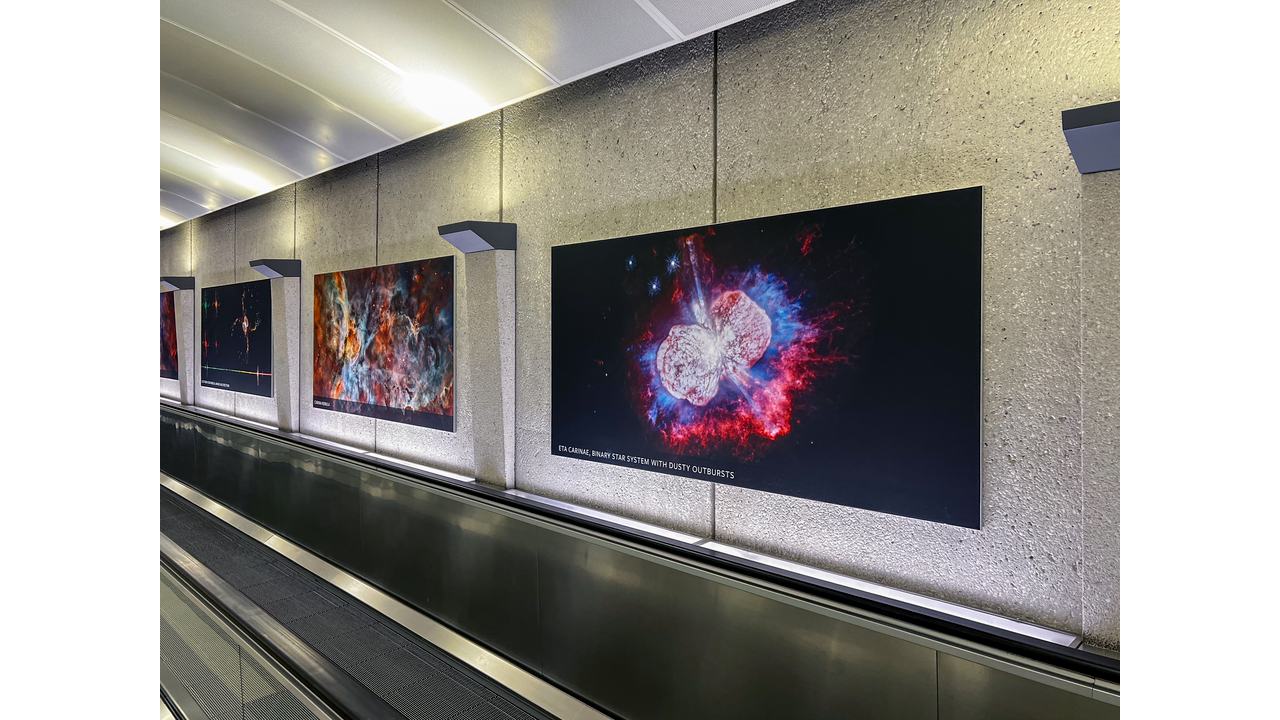 Closeup of Dulles airport hallway shows a large-scale Hubble Space Telescope image on the right, which takes up half of the view. It shows Eta Carinae, two roughly spherical white blobs of gas are connected by a bridge of material and look somewhat like an hourglass on its side. The object is set against the black background of space. To the left in the same hall are several additional large-scale Hubble images, each showing unique, colorful cosmic objects.