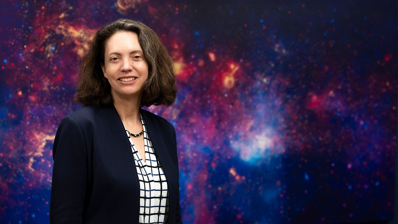 A photograph of a woman with shoulder-length dark hair wearing a dark blazer, checkered shirt, and necklace against a colorful background image of a nebula.
