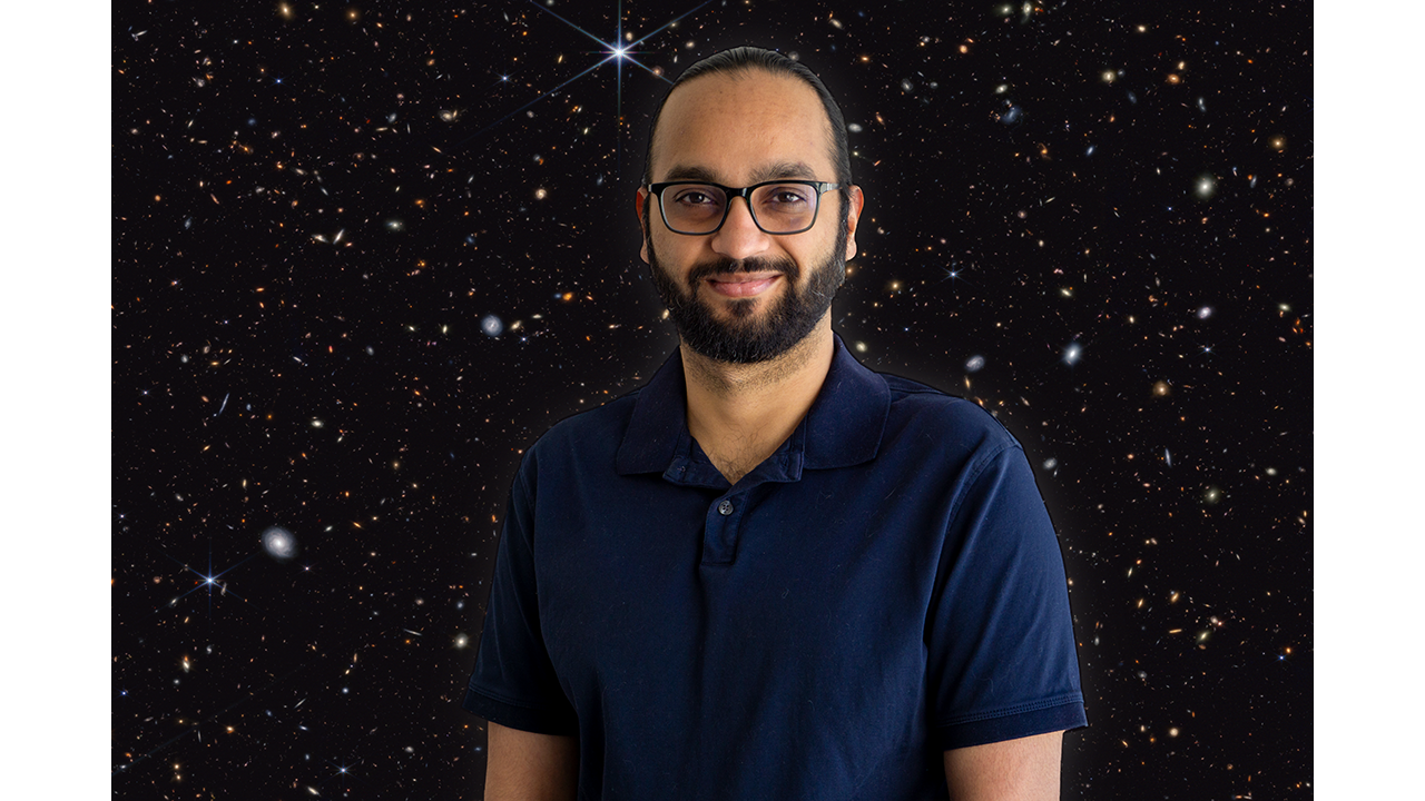 Gentleman with black hair, mustache, beard, and glasses, and wearing a dark blue, short-sleeved shirt, against a dark sky field featuring a multicolored sea of galaxies.